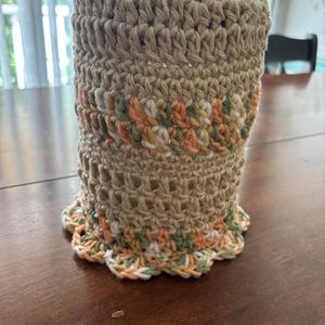 May include: A cylindrical crochet cosy featuring a solid tan yarn base with horizontal bands of variegated yarn in shades of peach, sage green, and white. The bottom edge is finished with a decorative scalloped border in the same variegated yarn.