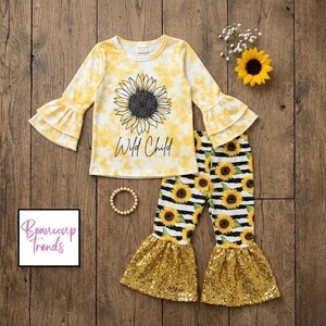May include: A yellow and white tie-dye long-sleeve shirt with a sunflower graphic and the text "Wild Child". The set includes matching pants with sunflower and stripe patterns, and gold sequin bell bottoms. A wooden beaded bracelet is also included.