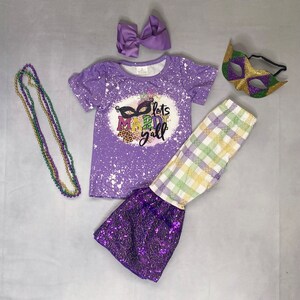May include: A purple Mardi Gras outfit set with a matching bow, mask, and beaded necklace. The set includes a short-sleeved shirt with the text "lets Mardi y'all", plaid pants, and sequin bell bottoms.