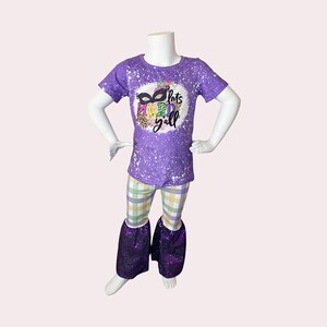 May include: A purple t-shirt and pants set on a mannequin. The t-shirt has a white splatter design and a graphic that says "lets MAREDI y'all" with a mask. The pants are plaid with purple sequin bell bottoms.