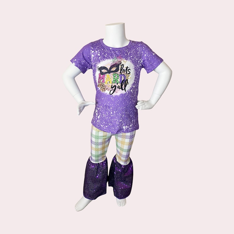 May include: A purple t-shirt and pants set on a mannequin. The t-shirt has a white splatter design and a graphic that says "lets MAREDI y'all" with a mask. The pants are plaid with purple sequin bell bottoms.