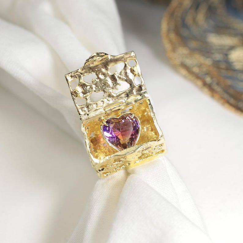 Op de afbeelding: Een goudkleurige, open doosvormige ring met een hartvormige, paarse edelsteen. De ring heeft een sierlijk, getextureerd ontwerp en is geplaatst op een witte doek. De edelsteen heeft een verloop van paarse en roze tinten.