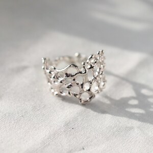 May include: A silver ring with a delicate, intricate design resembling coral or a honeycomb. The ring is made of a single piece of metal and has a unique, organic shape.