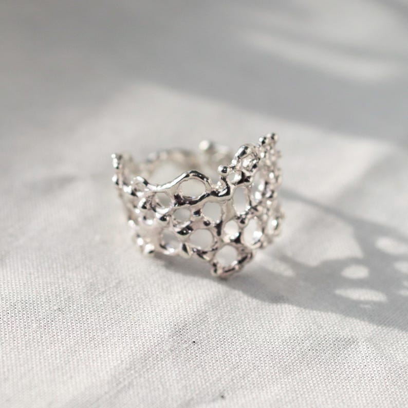 May include: A silver ring with a delicate, intricate design resembling coral or a honeycomb. The ring is made of a single piece of metal and has a unique, organic shape.