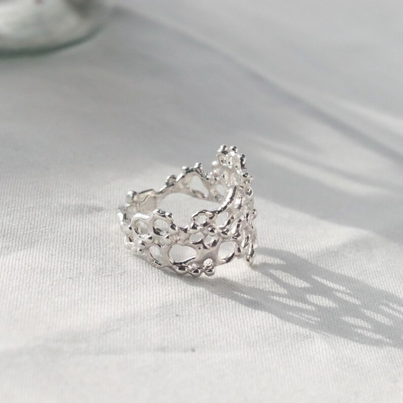 May include: A silver ring with a delicate, intricate design resembling coral. The ring is open and has a textured, organic look.