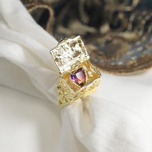 Op de afbeelding: Een goudkleurige ring met een getextureerde band en een scharnierende bovenkant. De bovenkant opent om een hartvormige edelsteen met paarse en roze tinten te onthullen. De ring ligt op een witte doek.
