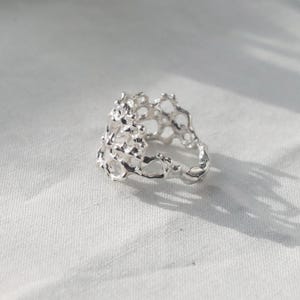 May include: A silver ring with a unique, organic, and textured design. The ring is made of a delicate, openwork pattern that resembles a coral reef or a honeycomb.