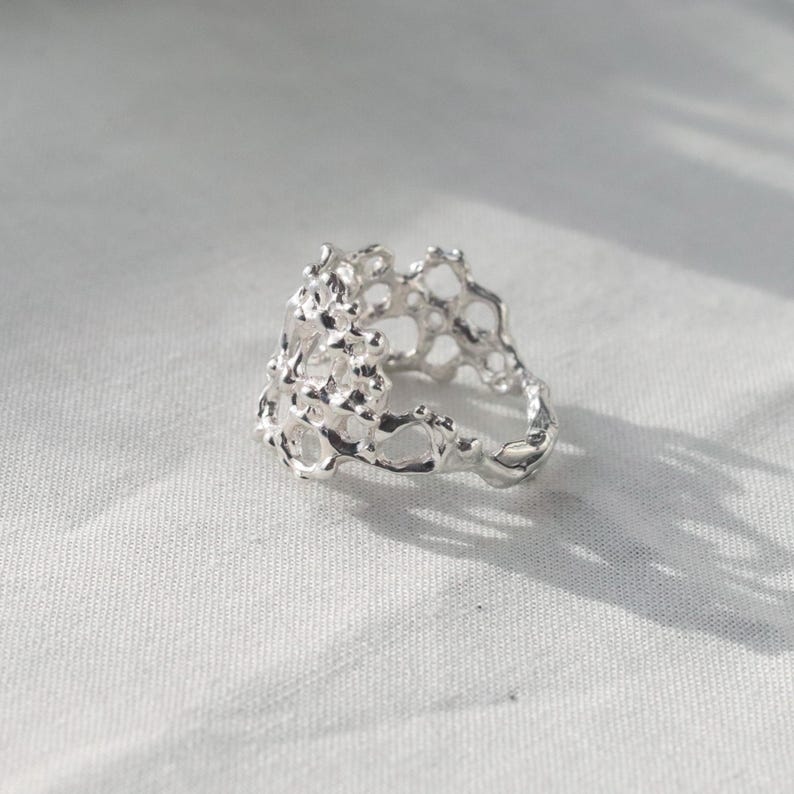 May include: A silver ring with a unique, organic, and textured design. The ring is made of a delicate, openwork pattern that resembles a coral reef or a honeycomb.