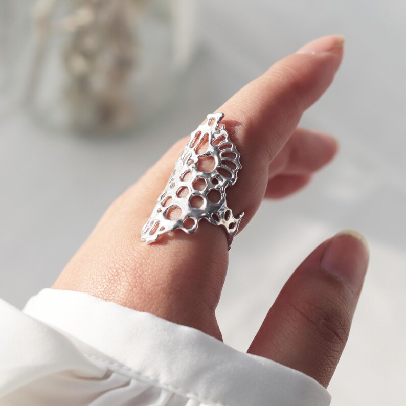 May include: A silver ring with a delicate, intricate design resembling a honeycomb or a floral pattern. The ring is worn on a finger and appears to be a statement piece.