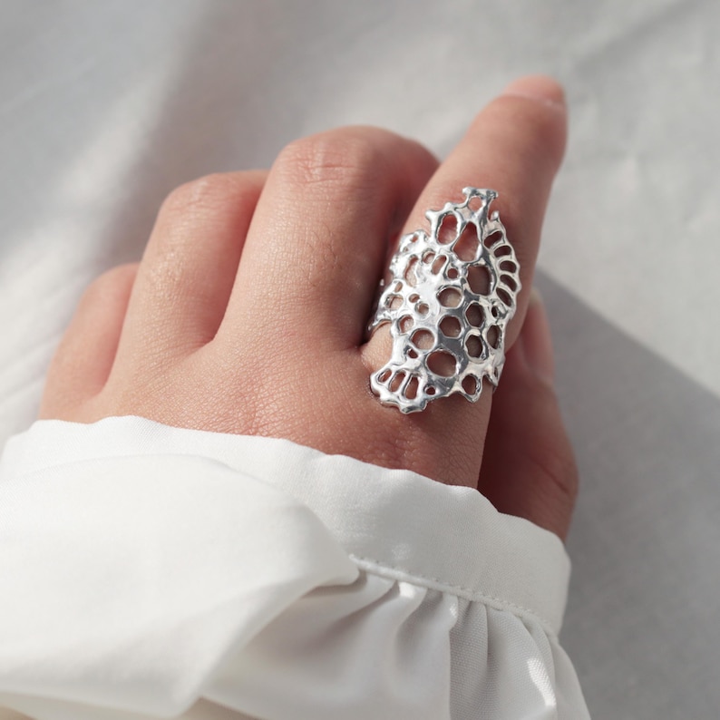May include: A silver ring with a delicate, intricate design resembling a piece of coral. The ring is large and has a unique, organic shape.