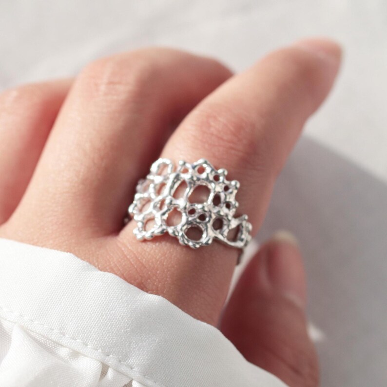 May include: A silver ring with a delicate, intricate design resembling a coral reef. The ring is shown on a finger.