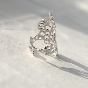 May include: A silver ring with a delicate, intricate design resembling a honeycomb or a coral reef. The ring is open and has a wide band.