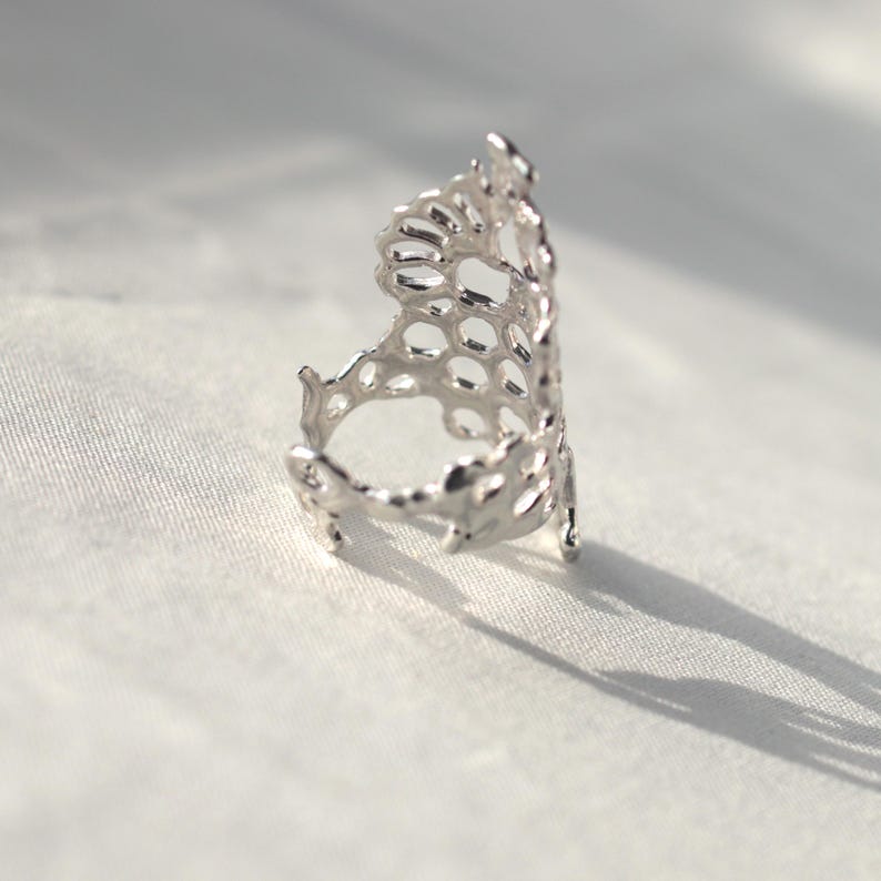 May include: A silver ring with a delicate, intricate design resembling a honeycomb or a coral reef. The ring is open and has a wide band.