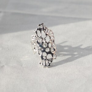 May include: A silver ring with a delicate, intricate design resembling a honeycomb or coral. The ring is made of a single piece of metal and has a unique, organic shape.