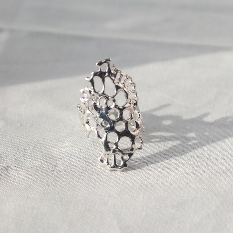 May include: A silver ring with a delicate, intricate design resembling a honeycomb or coral. The ring is made of a single piece of metal and has a unique, organic shape.