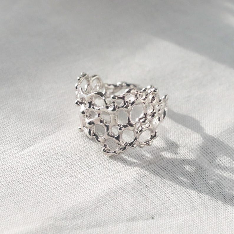 May include: A silver ring with a delicate, intricate design that resembles coral or a honeycomb. The ring is made of a single piece of metal and has a smooth, polished finish.