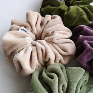 May include: A selection of velvet scrunchies in shades of beige, olive green, and purple. Each scrunchie has a small white tag with black text. The fabric appears soft and plush, with a luxurious texture.