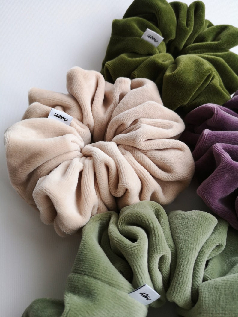 May include: A selection of velvet scrunchies in shades of beige, olive green, and purple. Each scrunchie has a small white tag with black text. The fabric appears soft and plush, with a luxurious texture.