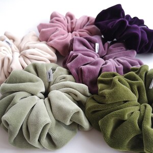 May include: A selection of large scrunchies in shades of beige, pink, purple, and green. The scrunchies are made from a soft, velvet fabric and have a small tag attached. They are arranged in a circular display, showcasing their colours.