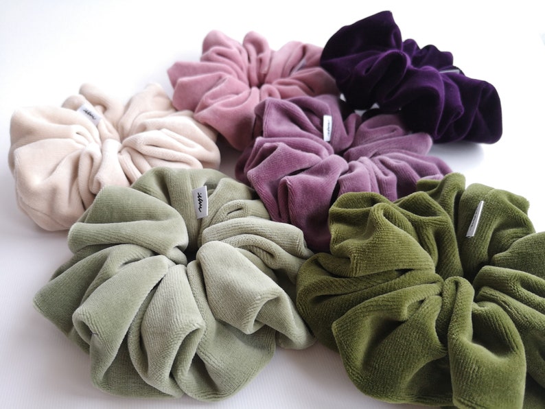 May include: A selection of large scrunchies in shades of beige, pink, purple, and green. The scrunchies are made from a soft, velvet fabric and have a small tag attached. They are arranged in a circular display, showcasing their colours.