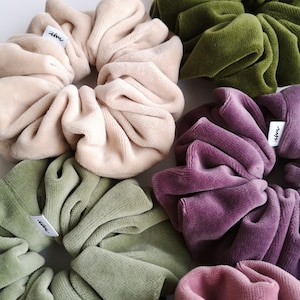 May include: An assortment of large velvet scrunchies in shades of beige, olive, purple, and pink. Each scrunchie features a small branded tag. These soft hair accessories are made from a plush material.