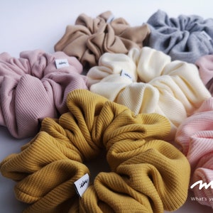 May include: A collection of colourful, ribbed hair scrunchies in various shades of pink, yellow, beige, and grey. The scrunchies are made of soft, stretchy fabric and have a simple, minimalist design. Each scrunchie has a small white tag with the word "mini" printed on it.