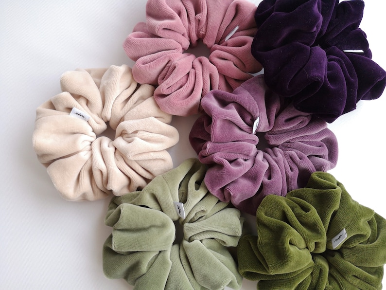 May include: A selection of large scrunchies in shades of beige, pink, purple, and green. Each scrunchie is made from a soft, velvety material and has a small white label. The scrunchies are arranged in a circular pattern.