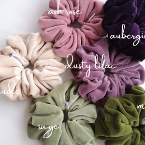 May include: An assortment of large velvet scrunchies in shades such as nude, ash rose, dusty lilac, aubergine, sage, and moss. Each scrunchie has a soft, velvety texture and a small white tag. The scrunchies are displayed on a white background.