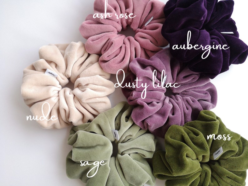 May include: An assortment of large velvet scrunchies in shades such as nude, ash rose, dusty lilac, aubergine, sage, and moss. Each scrunchie has a soft, velvety texture and a small white tag. The scrunchies are displayed on a white background.