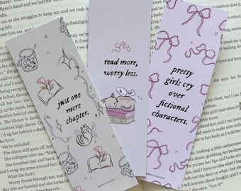 May include: Three decorative bookmarks with whimsical illustrations and text. One says "just one more chapter," another "read more, worry less," and the third "pretty girls cry over fictional characters." The bookmarks are on a book.