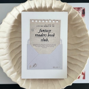 May include: A brown and white card with the text "all access pass LIFETIME MEMBER OF THE fantasy readers book club." The card is in the shape of an envelope with a small illustration of a cat reading a book in the lower left corner.