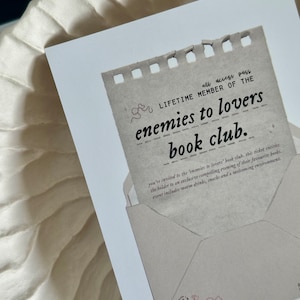 May include: A brown paper card with a torn edge, featuring the text "all access pass LIFETIME MEMBER OF THE enemies to lovers book club." The card is decorated with a small illustration of a bear holding a flower and a bow.
