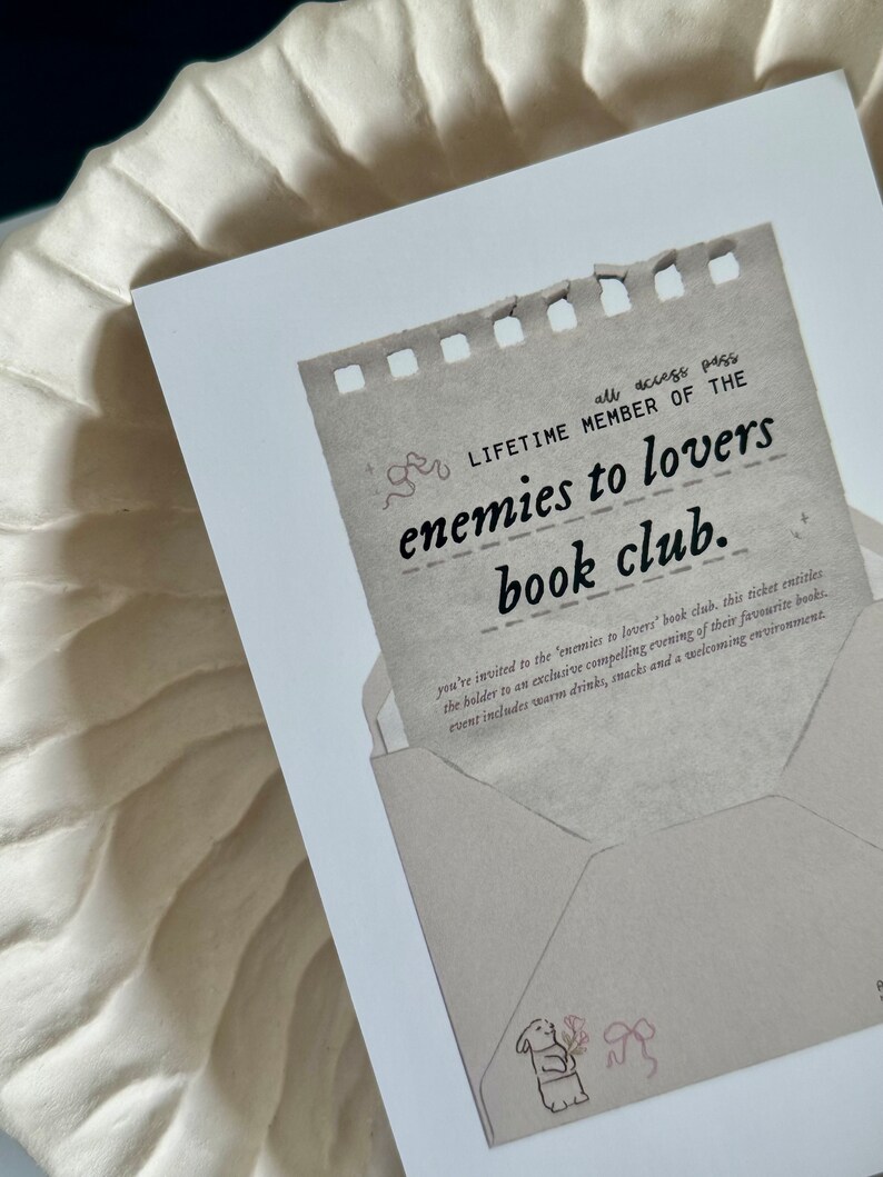 May include: A brown paper card with a torn edge, featuring the text "all access pass LIFETIME MEMBER OF THE enemies to lovers book club." The card is decorated with a small illustration of a bear holding a flower and a bow.