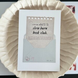 May include: A white card with a brown paper insert that reads "all access pass LIFETIME MEMBER OF THE slow burn book club." The card has a blue envelope with a pink ribbon and a pink bow on it. The card also has the text "ADMIT ONE" on it.