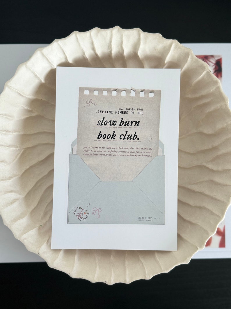 May include: A white card with a brown paper insert that reads "all access pass LIFETIME MEMBER OF THE slow burn book club." The card has a blue envelope with a pink ribbon and a pink bow on it. The card also has the text "ADMIT ONE" on it.
