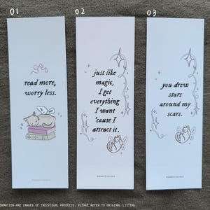 Op de afbeelding: Drie witte bladwijzers met roze en grijze illustraties en tekst. De eerste bladwijzer toont een kat die slaapt op een stapel boeken met de tekst "lees meer, maak je minder zorgen." De tweede bladwijzer toont een kat met vleugels en de tekst "net als magie, krijg ik alles wat ik wil omdat ik het aantrek." De derde bladwijzer toont een kat met vleugels en de tekst "je hebt sterren getekend rond mijn littekens."