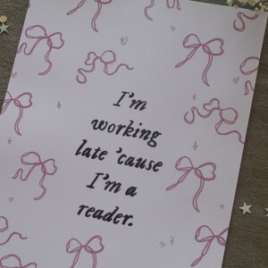 Op de afbeelding: Een roze printbare kaart met een quote die luidt: "Ik werk laat omdat ik een lezer ben." De achtergrond is versierd met roze strikken.