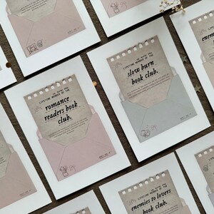 May include: A set of ten white framed prints with brown paper envelopes inside. Each envelope has a different book club theme written on it, such as "cozy readers book club" and "enemies to lovers book club".