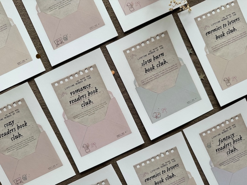 May include: A set of ten white framed prints with brown paper envelopes inside. Each envelope has a different book club theme written on it, such as "cozy readers book club" and "enemies to lovers book club".