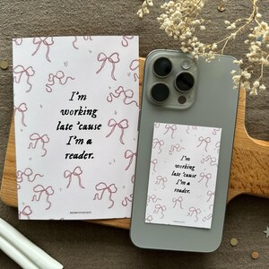 Op de afbeelding: Twee witte kaarten met roze strikken en de tekst "Ik werk laat omdat ik een lezer ben". Een kaart zit op een witte telefoonhoes met een zwarte camera lens.