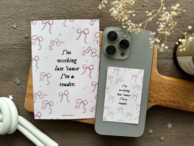 Op de afbeelding: Twee witte kaarten met roze strikken en de tekst "Ik werk laat omdat ik een lezer ben". Een kaart zit op een witte telefoonhoes met een zwarte camera lens.