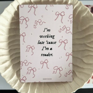 Op de afbeelding: Een roze en witte kaart met een quote die luidt: "Ik werk laat omdat ik een lezer ben." De kaart is versierd met roze strikken.