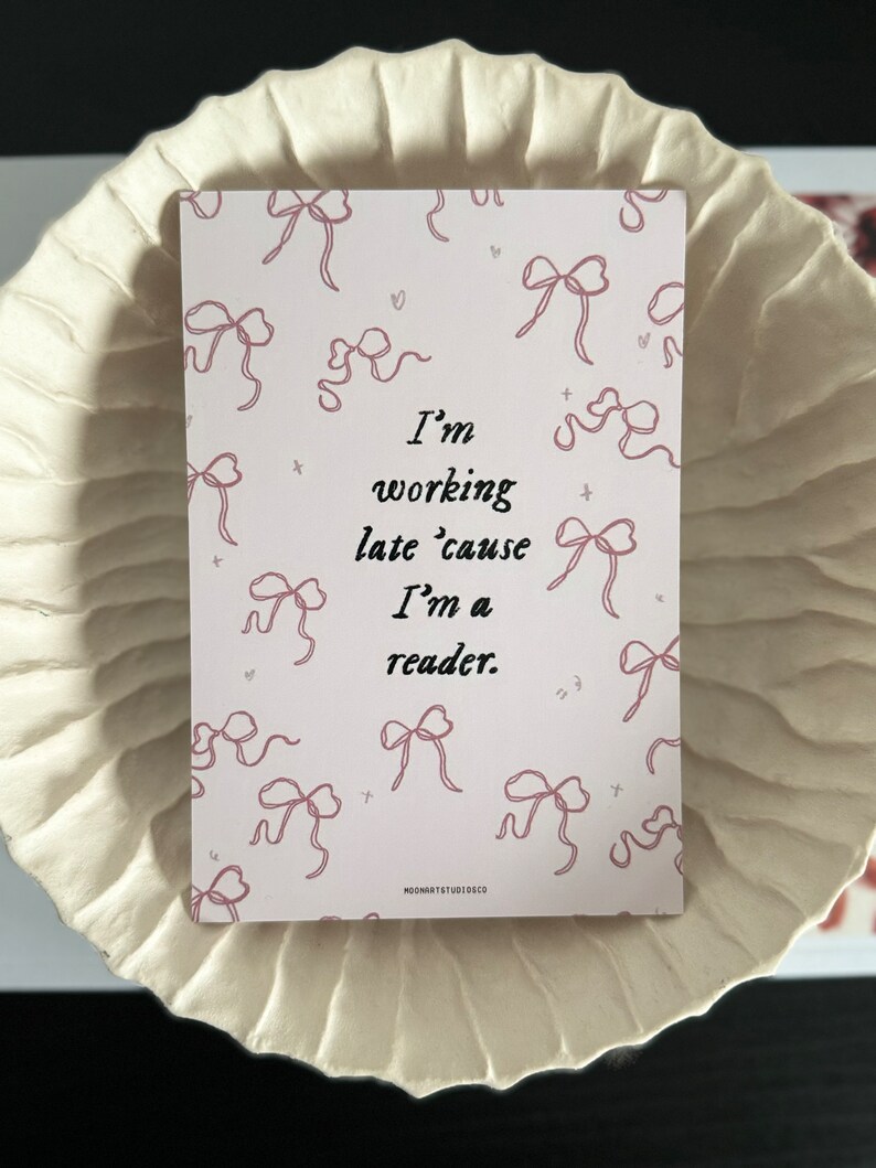 Op de afbeelding: Een roze en witte kaart met een quote die luidt: "Ik werk laat omdat ik een lezer ben." De kaart is versierd met roze strikken.
