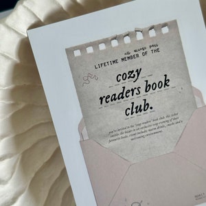 May include: A vintage-style ticket with a torn edge, reading "all access pass LIFETIME MEMBER OF THE cozy readers book club." The ticket is in a pink envelope with a hand-drawn illustration of a cat curled up with a mug.