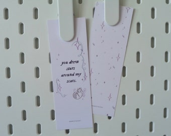 May include: Two pink bookmarks with a whimsical design. One bookmark features a hand-drawn illustration of a cat with wings and the text "you drew stars around my scars." The other bookmark has a repeating pattern of stars.