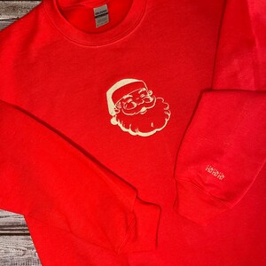 Op de afbeelding: Rood sweatshirt met een wit geborduurd beeld van de kerstman op de voorkant en de tekst "HO HO HO" op de linkermouw.