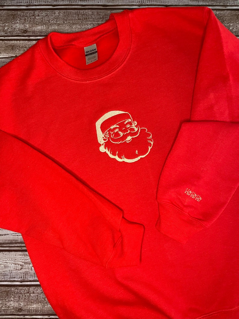 Op de afbeelding: Rood sweatshirt met een wit geborduurd beeld van de kerstman op de voorkant en de tekst "HO HO HO" op de linkermouw.