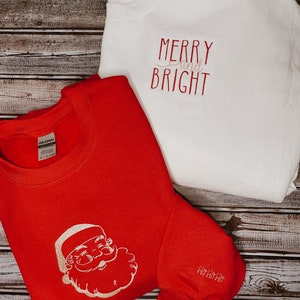 Op de afbeelding: Twee rode en witte kersttruien. E&eacute;n trui heeft een witte kerstman-afbeelding en de tekst "Ho Ho Ho" op de mouw. De andere trui heeft de tekst "Merry and Bright" in wit.