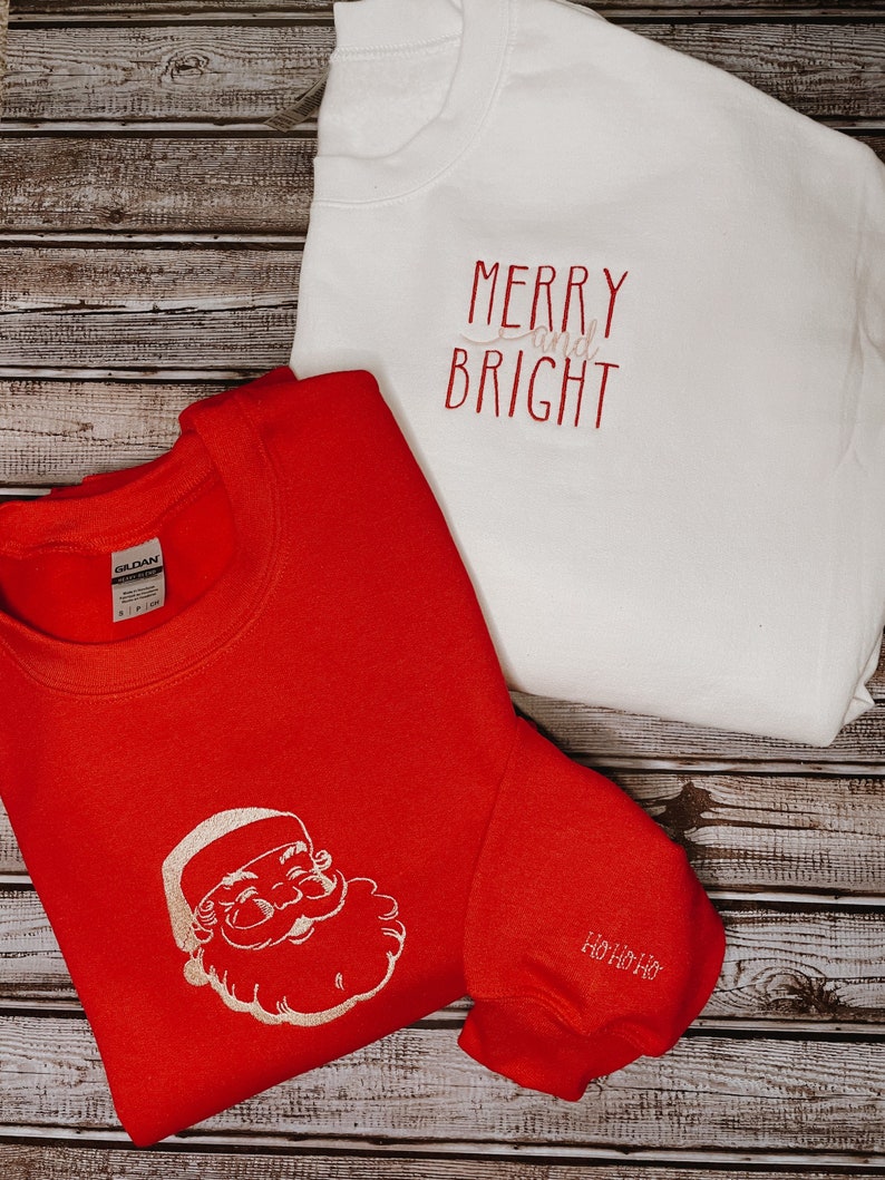 Op de afbeelding: Twee rode en witte kersttruien. E&eacute;n trui heeft een witte kerstman-afbeelding en de tekst "Ho Ho Ho" op de mouw. De andere trui heeft de tekst "Merry and Bright" in wit.