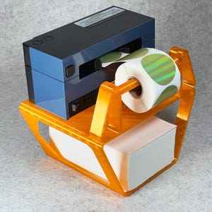May include: A blue label printer with a roll of iridescent stickers and a stack of white paper in an orange plastic stand.